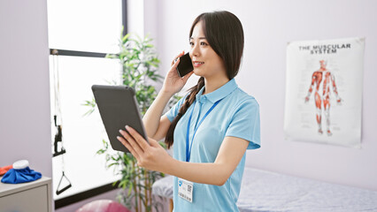 A young asian woman healthcare professional talks on a phone while holding a tablet in a clinic...