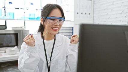 Excited asian woman scientist wearing lab coat and safety glasses in laboratory celebrates success