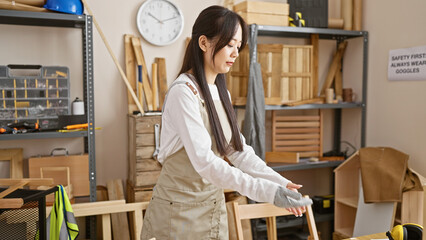 A young asian woman in a carpentry workshop wearing gloves and an apron stands surrounded by wood...