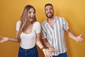 Young couple standing over yellow background smiling cheerful with open arms as friendly welcome, positive and confident greetings