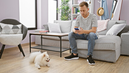 Engrossed young caucasian man sitting on living room sofa, texting on his smartphone while his...