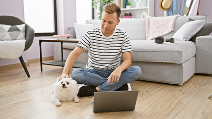 Focused young caucasian man sitting with his loving pet dog on the floor, intently using his laptop...