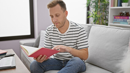 Young caucasian man reading book sitting on sofa at home