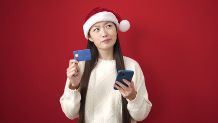 Young chinese woman wearing christmas hat using smartphone and credit card over isolated red background