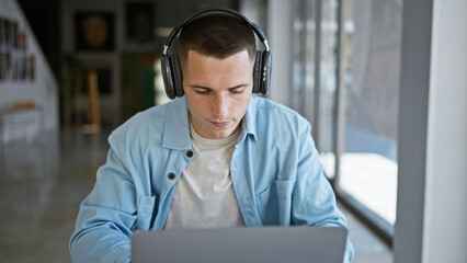 A focused young hispanic man in headphones works on his laptop at a modern university library.