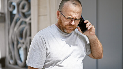 Handsome middle-aged caucasian man standing on sunlit urban street, engrossed in serious phone...