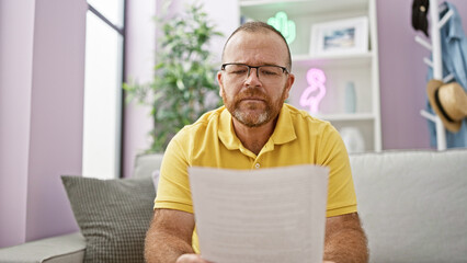 Fototapeta premium Upset, handsome adult male studying indoors, serious caucasian man reading a complex document while sitting on his living room sofa at home