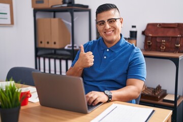 Young hispanic man working at the office with laptop doing happy thumbs up gesture with hand. approving expression looking at the camera showing success.