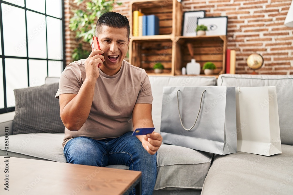 Sticker hispanic young man doing payment with credit card on the phone winking looking at the camera with se