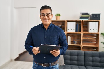 Young latin man business worker smiling confident holding clipboard at office