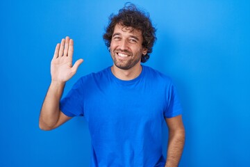 Naklejka premium Hispanic young man standing over blue background waiving saying hello happy and smiling, friendly welcome gesture