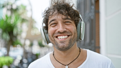 A cheerful young hispanic man wearing headphones poses for a portrait on a sunlit urban street.