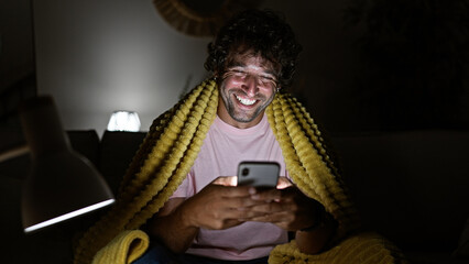 A cheerful young hispanic man enjoys browsing his smartphone at home, wrapped in a cozy yellow...