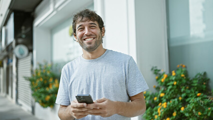 Happy young man, sporting a beard and blond hair, enjoying his time outdoor in the city, confidently smiling while typing a message on his smartphone screen - obrazy, fototapety, plakaty
