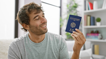 Happy young man, sitting casually on his living room sofa, smiling, holding his passport, thrilled...