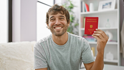 Fototapeta premium Casual joy! young, happy guy with blond beard, confidently smiling, holding his austrian passport, enjoying a quiet moment on the sofa at home. the perfect adult, patriotic vacation mood!