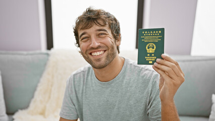 Cheerful young man, casually sitting on the living room sofa, joyfully holds his macao passport...