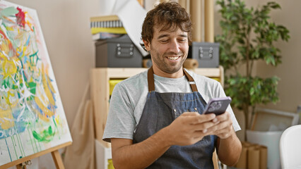 Cheerful young hispanic artist man, blond beard and apron, smiling while typing a message on his...