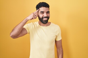 Hispanic man with beard standing over yellow background smiling pointing to head with one finger, great idea or thought, good memory