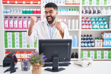 Hispanic man with beard working at pharmacy drugstore very happy and excited doing winner gesture with arms raised, smiling and screaming for success. celebration concept.