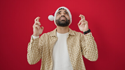 Young hispanic man wearing christmas hat crossing fingers for luck over isolated red background