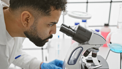A focused young adult man with a beard wearing lab coat and gloves examines samples through a...