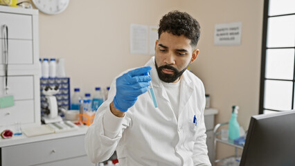 Naklejka na ściany i meble A focused latino man in a lab coat examines a test tube in a modern laboratory environment.