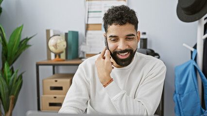 Fototapeta premium A smiling young hispanic man with a beard talking on a mobile phone in a modern office setting