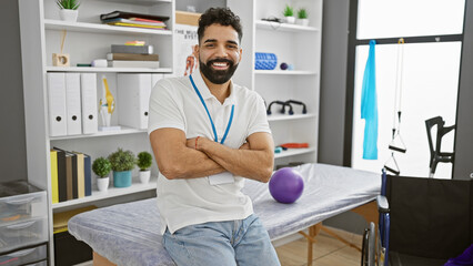 Young hispanic man with beard smiling confidently while standing arms crossed in a modern rehabilitation clinic interior - Powered by Adobe