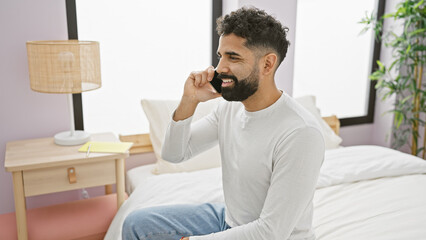 Smiling bearded man talking on phone in a cozy bedroom setting, conveying casual relaxation and comfortable home life