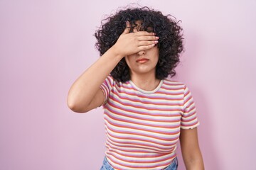 Young middle east woman standing over pink background covering eyes with hand, looking serious and...