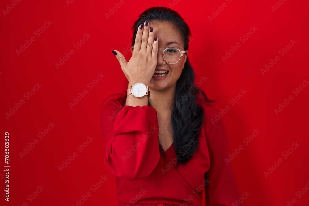 Poster asian young woman standing over red background covering one eye with hand, confident smile on face a