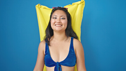 Young chinese woman tourist wearing bikini smiling over float mattress over isolated blue background