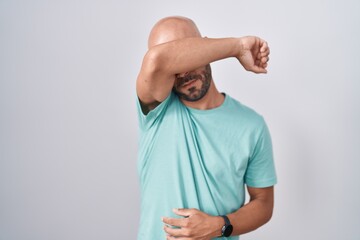 Middle age bald man standing over white background covering eyes with arm, looking serious and sad....