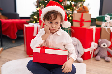 Fototapeta na wymiar Adorable hispanic boy holding christmas gift sitting on floor with unhappy expression at home