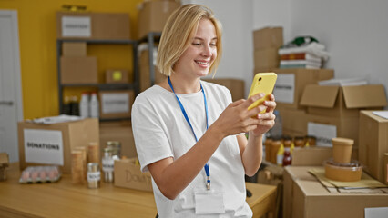 Young blonde woman volunteer using smartphone smiling at charity center