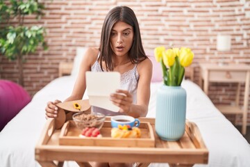 Brunette young woman eating breakfast in the bed reading a letter afraid and shocked with surprise...