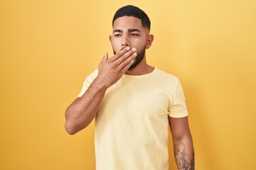 Young hispanic man standing over yellow background bored yawning tired covering mouth with hand. restless and sleepiness.