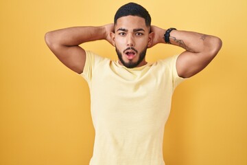 Young hispanic man standing over yellow background crazy and scared with hands on head, afraid and surprised of shock with open mouth