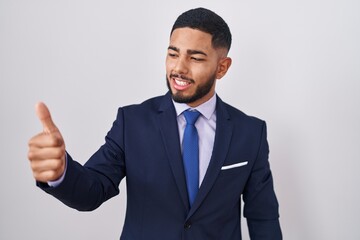 Young hispanic man wearing business suit and tie looking proud, smiling doing thumbs up gesture to...