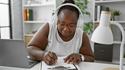 Smiling african american woman boss taking notes, working online on laptop in office. plus size worker with braids, headphones conquering business, exudes professional success indoors