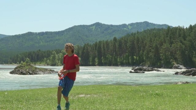 Slow motion. Young Boy throwing a red frisbee disk.  Young boy playing on a mountain river bank. Cute boy playing with frisbee outdoor on sunny day. Against the backdrop of mountains, on the coast 