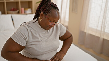 African american woman, sitting in bed, suffering intense menstrual pain, battling abdomen ache in...