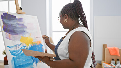 Focused african american woman artist, with braids and glasses, seriously drawing on canvas at...