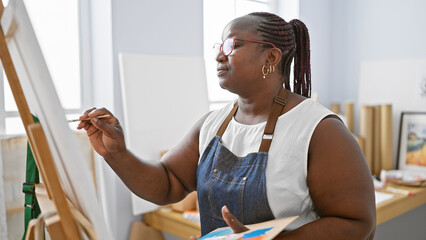 Focused african american woman artist, with braids and glasses, seriously drawing on canvas at...