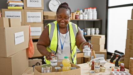 Smiling african american woman volunteering, packing food into cardboard box for charity donation...