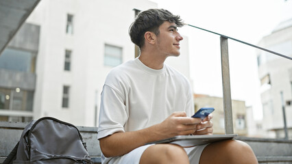 Sunlit young hispanic university student, joyfully texting on his smartphone while sitting on city...