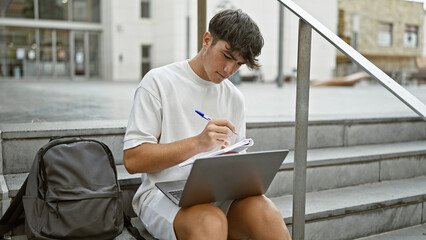 Cool hispanic teenager relaxes on university stairs, seriously concentrated on taking notes on his laptop