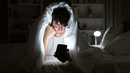 Young hispanic teenager seriously engrossed in texting on his smartphone, lying on a comfy bed...