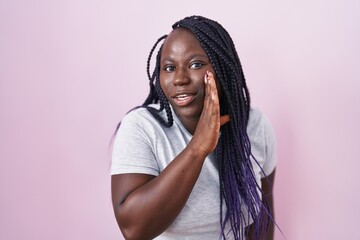 Young african woman standing over pink background hand on mouth telling secret rumor, whispering...
